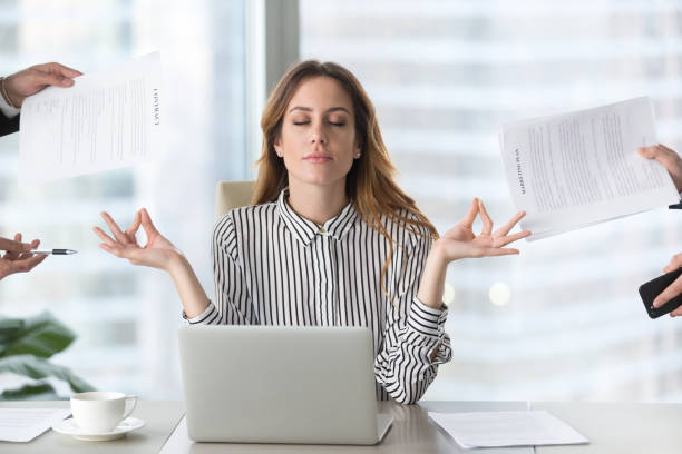 Calm female executive meditating at her office desk, eyes closed and hands in a mudra, taking a mindful break from work to reduce stress and find mental balance.